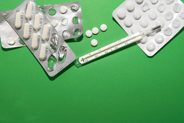 Packs of pills and a thermometer lie on a green background, next to them are several tablets...