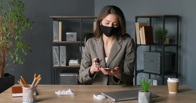 Stylish Caucasian woman in mask sitting at table and wiping smartphone with napkin in office. Coronavirus pandemic protection. Female cleaning, sterilizing and sanitizing mobile phone with sanitizer