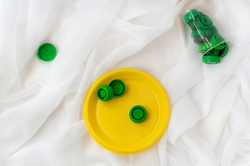 Heap of plastic green, yellow bottle caps on disposable plate and cups on white cloth. Save earth, go green, recycling concept. Creative flat lay, designer background.