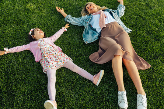 Above view image of happy mom lying on the green grass with her cute little girl in the park. Cheerful young woman relaxing with her child, enjoying the time together. Mother and daughter shares love