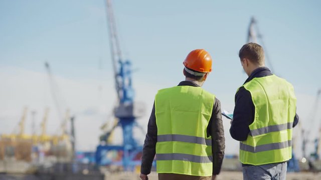 Back view medium shot of two shipbuilding engineers examining vessels under construction and discussing plans at dockside, copy space to left