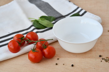 White empty bowl near a sprig of cherry tomatoes and baby spinach, with kitchen towel, on wooden table, front view