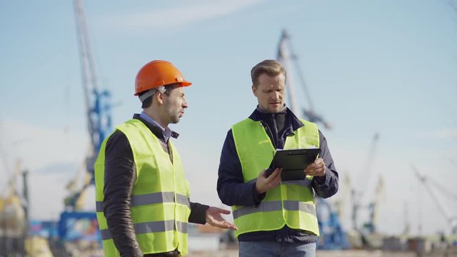 Medium shot of two engineers walking at shipbuilding yard, observing constructions of vessels and discussing plans. Middle aged specialist with clipboard speaking into walkie-talkie