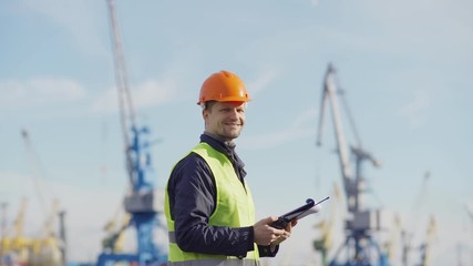 Side view medium shot of middle aged marine engineer in orange helmet and with clipboard speaking into walkie-talkie at dockside, then turning his head, looking at camera and smiling - Powered by Adobe