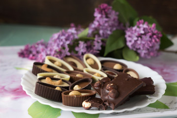 Bouquet of lilac flowers and chocolate candies with filling and nuts, chocolates with nuts on a plate, background. Sweets made of black, milk and white chocolate with hazelnuts, almonds and cashews