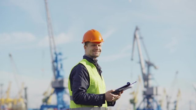 Side view medium shot of middle aged marine engineer in orange helmet and with clipboard speaking into walkie-talkie at dockside, then turning his head, looking at camera and smiling