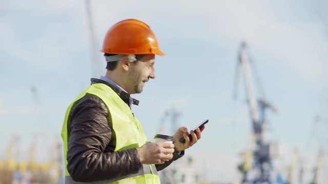 Side view tracking shot of middle aged port engineer in helmet and safety vest walking at dockyard, drinking coffee, reading text message on cell phone and looking around