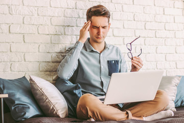 Stressed young hipster man freelancer feeling headache while working with laptop at home....