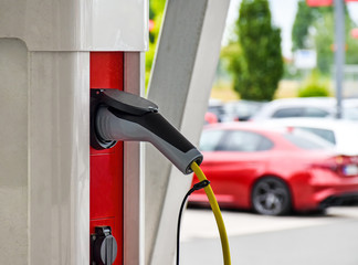 A charging cable for electric vehicles is plugged into a red-white charging station, cars are visible in the background.