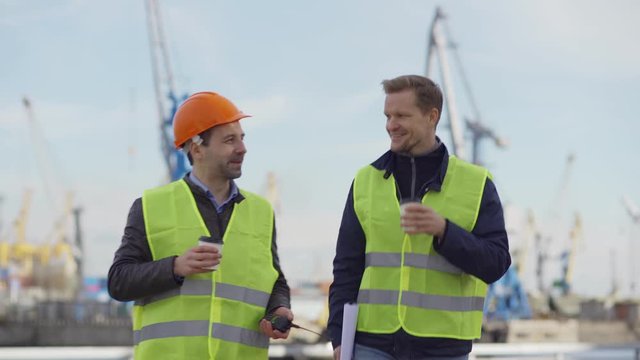 Medium shot of two smiling middle aged port engineers walking at dockyard, drinking coffee and talking at break