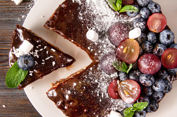 piece of cake, chocolate cake with fresh berries, blueberries, grapes, powdered sugar and mint on a white plate on a wooden , close up,  top view