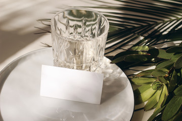 Summer tropical stationery still life scene. Glass of water, palms leaves, leucadendron flower. Beige table background in sunlight. Blank business, greeting card, invitation mockup scene. Long shadows