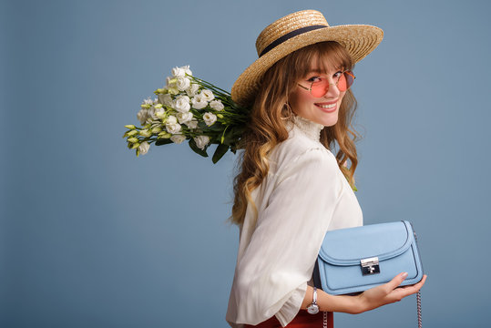 Fashion portrait of elegant happy smiling model wearing trendy spring outfit: white blouse, straw hat, holding small stylish faux leather bag, posing on blue background. Copy, empty space for text