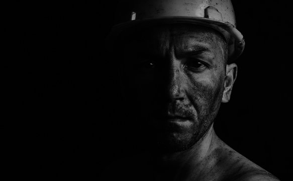 Dirty coal miner in a yellow hard hat on a dark background in a black and white photo.