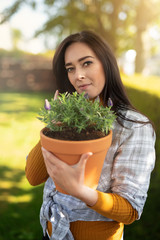 Beautiful dark haired smiling woman holding pot with plant while standing in the garden