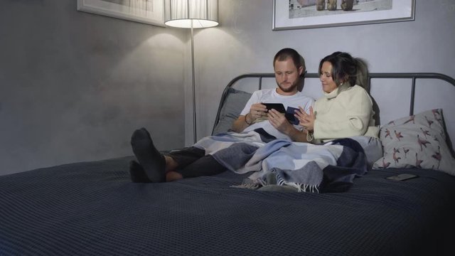 Waist-up arc shot of young Caucasian husband and wife in pyjamas sitting up silently in bed at night, leaning on headboard, holding smartphones and looking completely immersed