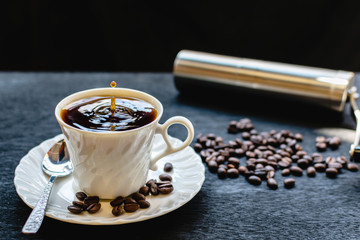 A white cup with a splash of drops coffee in the morning, The water surface on the glass fluttering, Antique coffee grinder using hand crank and dark roasted coffee beans on black background.