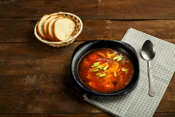 Black dish of italian tomato soup with potatoes and mushrooms on a wooden table on a napkin and a spoon and bread.