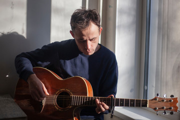 Obraz premium Guy with a guitar near the window. The guy in the morning light in the kitchen. The man is rehearsing music. Playing an acoustic instrument. Guitar lesson.