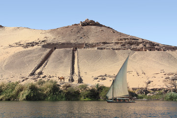 An Egyptian sailboat photographed from the great river Nile in front of a large rocky and sandy hill in the afternoon in the sunshine. There is a temple on the mountain and a camel on the bank below.
