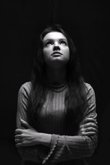 Black and white portrait of a young girl looking up girl with long hair.  Woman prying. Top light. Dramatic girl on a dark background. Selective lighting. Concept of religion, hope and prayers.