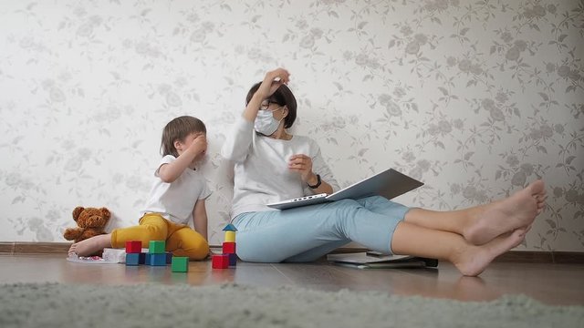 Mom and son sit at home quarantine because of coronavirus COVID19. Mother works remotely with laptop, son plays with toy blocks. Self isolation at home.