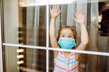 Little girl wearing a medical mask in quarantine staying behind big window with her hands up. Coronavirus quarantine concept