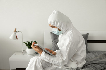 Man in protective white suit and medical mask is working from home on a bed with a tablet because of coronavirus epidemic. Remote work during pandemic. Stay home during COVID-19 quarantine concept.