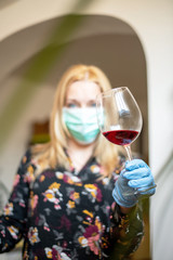 girl with golden hair in a surgical mask and protective gloves holds a glass of red wine, vertical position