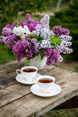 Lilac spring flowers in vase on wooden background. Tea coffee cup on table during breakfast time