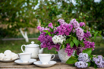Lilac spring flowers in vase on wooden background. Tea coffee cup on table during breakfast time