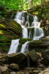 Mountain waterfall in a green forest among stones and trees. Cascade falls over mossy rocks