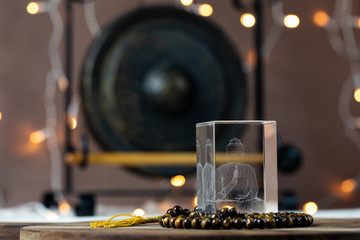 Teaching buddha statue in engraved crystal glass standing in tiger eye gemstone Mala prayer beads with a gong and bokeh lights in background