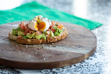 Healthy vegan vegetarian meal with chia and sesame seeds sprinkled over a runny poached egg, diced tomatoes and avocado on a cereal toasted bread slice, on a wooden plate over a table with green mat