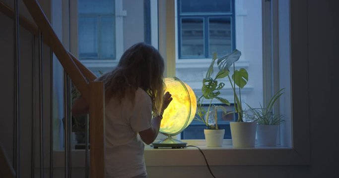 Young teenage girl in pyjamas, looks at map on lit up globe. Cinematic shot in concept of missed summer vacation, locked down at home during quarantine. Planning dreams and wanderlust adventures