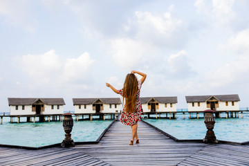 Back view of joyful cute attractive young happy long-hair girl with arms up standing on a wooden pier and enjoying ocean, summer breeze and sound of the waves during vacation. holiday travel concept.