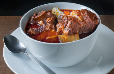 Mexican Food Concept, beef ribs soup in a white bowl, with a spicy salsa made of chile guajillo and other hot chilis and tomatoes with vegetables like zucchini, carrots, in a dark background.
