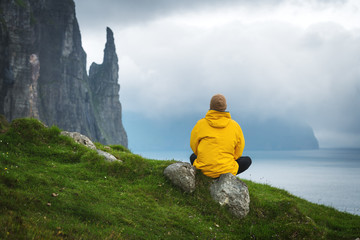 Tourist in yellow jacket looks at Witches Finger cliffs from Trollkonufingur viewpoint. Vagar island, Faroe Islands, Denmark.