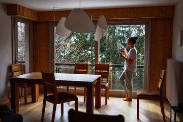 Young woman standing with cellphone and texting next to the window. Beautiful forest view outside. Wide shot of the apaetment. Stay home concept.