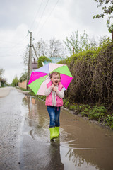 A beautiful little girl walks through puddles with a colorful umbrella and green rubber boots. Walk a cute girl in the fresh air after the rain.
