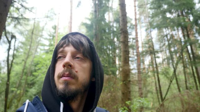 guy in a black hood walks through a pine forest in the afternoon, in spring and looks for a way out since he got lost and takes off himself