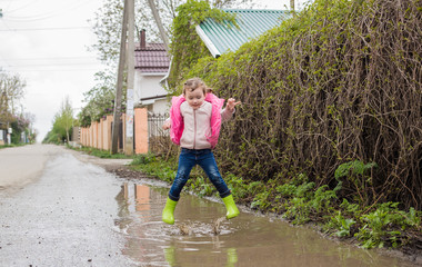 Fototapeta na wymiar A beautiful little girl in green rubber boots jumps in puddles. Shooting on the move.