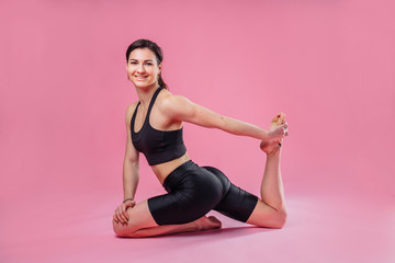 Sportive girl fitness trainer dressed in a black top and black leggings sitting on the floor on a pink background in the studio. Demonstrating leg muscle stretching exercise and smiling.