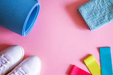 Fitness flat-lay with sneakers, blue mat, towel, elastic rubber bands on pink background