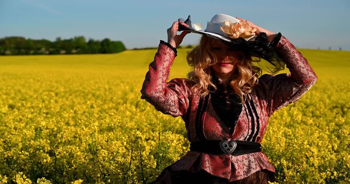 Blond noblewoman wearing large hat and historical costume standing in the yellow oilseed rape field 