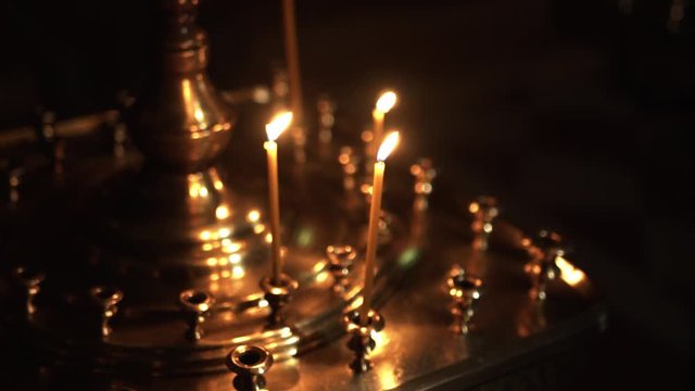 In a dark room there is a tall golden candlestick in which several bright candles burn and illuminate the gloomy room of the church, close-up, side view