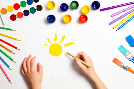 Kid hands painting yellow sun. Top view to white table with art supplies