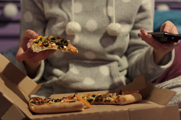 Woman eating pizza at home in her pajamas and watching tv. With Pizza slice on one hand and tv remote on the other