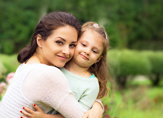 Fototapeta premium Beautiful mother hugging and kissing her cute small daughter on summer green grass background. Closeup