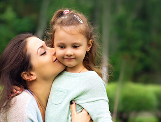 Beautiful mother hugging and kissing her cute small daughter on summer green grass background. Closeup